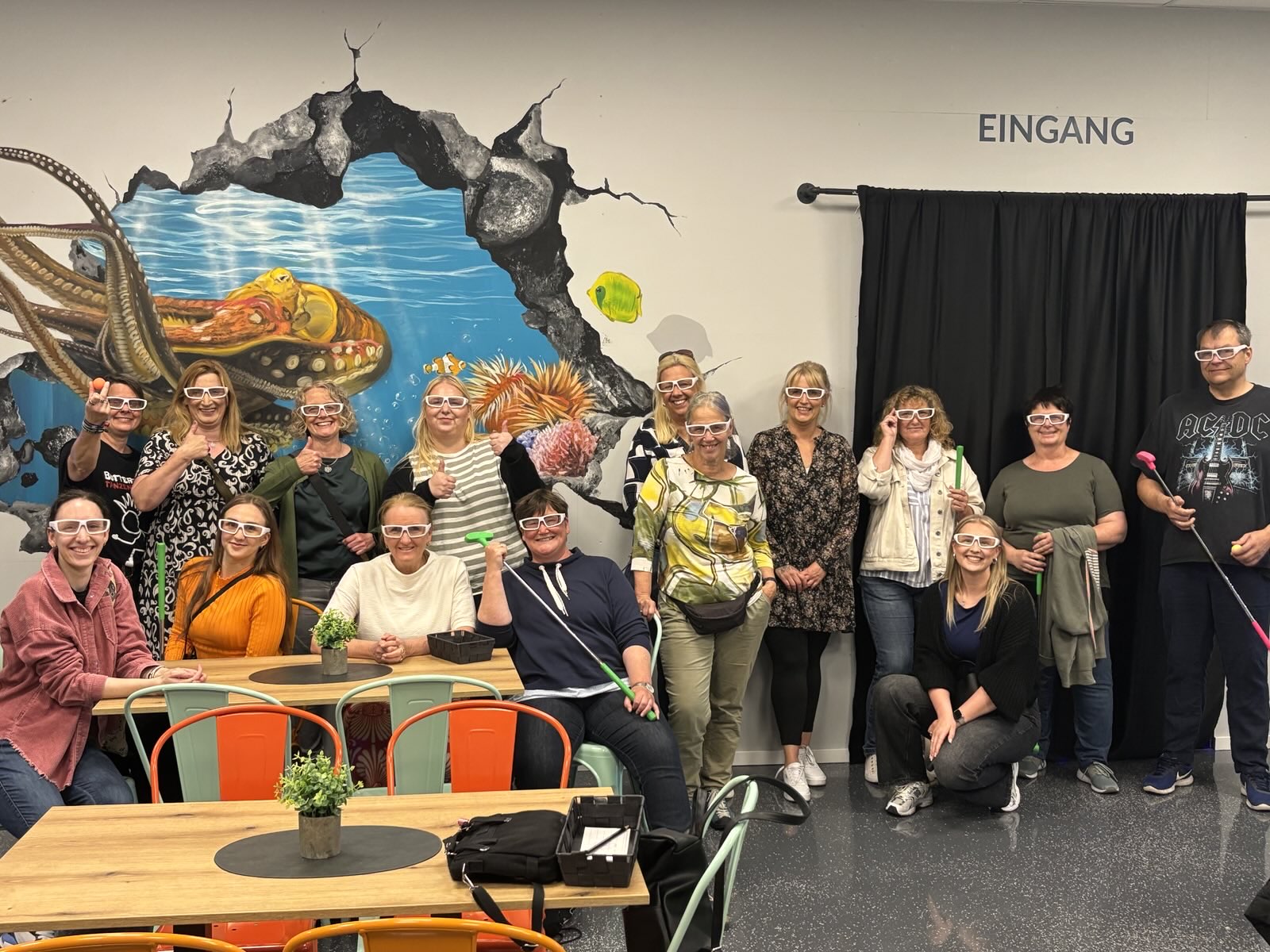 Foto vor dem Eingang des Schwarzlicht-Minigolf mit dem Team des Integrationsfachdienstes. Das Team lächelt fröhlich in die Kamara und trägt 3D-Brillen, Einzelne halten Golfschläger in der Hand. Eine Kollegin hält einen orangen Golfball ins Bild. Von der Gruppe von 15 Leuten stehen die meisten in einer Reihe, vier sitzen auf der linken Seite an einem Tisch, eine Person sitzt in der Hocke. Auf die weißen Wand ist ein Loch gezeichnet, in dem eine Krake ins Meer schwimmt.